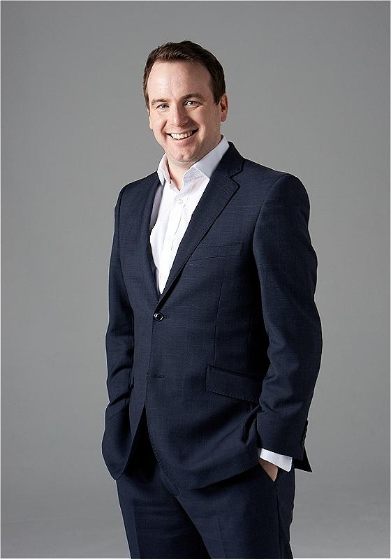 Matt Forde, wearing a navy suit with no tie, smiles to the camera. 