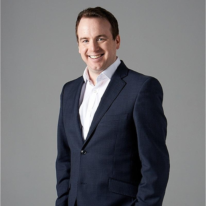 Matt Forde, wearing a navy suit with no tie, smiles to the camera. 