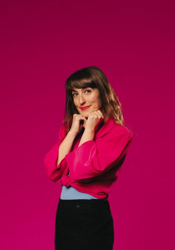 Woman stands in front of pink backdrop with her hands holding her chin and looking adorably at the camera 