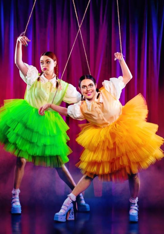 Two sisters stand in odd positions attached to puppet strings in neon tutu dresses and white shirts