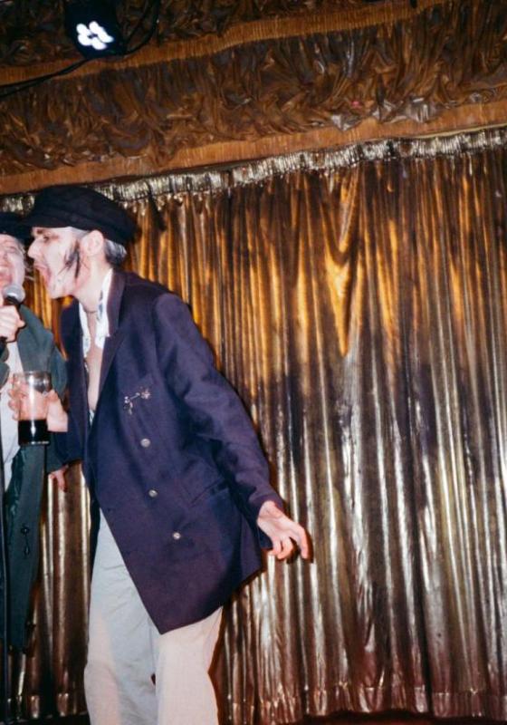 Two drag kings dressed as old men scream into a microphone on a stage with a gold curtain backdrop