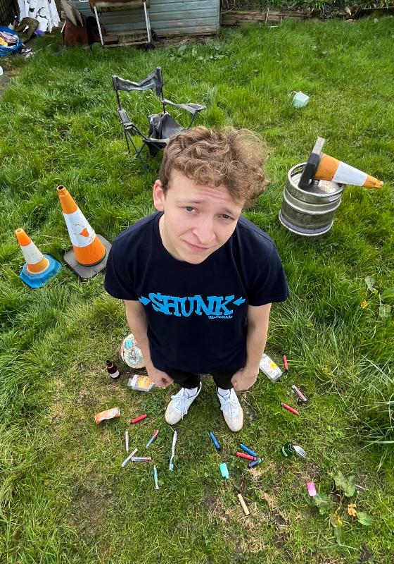 A young white man in his early twenties with light brown curly hair is standing in a very messy garden. He is looking up at the camera with a standoff-ish expression. He is wearing a dark blue shirt that says 'Shunk' on it in light blue letters. At his feet in the grass are about 15 used vapes of different colours and sizes, an old football and empty beer and spirit bottles. Just behind him on the left of the picture are two orange and white traffic cones. slightly above that is a tattered old garden chair,
