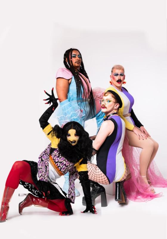 A studio shot of drag performers in costume; Bard the Beholder, Cyro, Flick and Carrot, all looking sassy and posing at different heights and levels. They are giving gender in different transient forms.