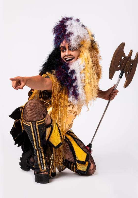 	An image of Oedipussi Rex, a white Drag Barbarian with a long beard and wig made out of tinsel in the colours of the Non-Binary Flag. He is Kneeling, his body facing right, while looking and pointing to something out of shot to the left. His expression is gleeful, and he is holding a large double edged axe in his right hand. He is also wearing golden ancient Greek style armour, complete with platform gladiator sandals.