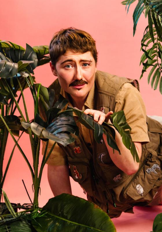Performer is dressed as explorer in beige crawling through a jungle backdrop of house plants against a pink background.