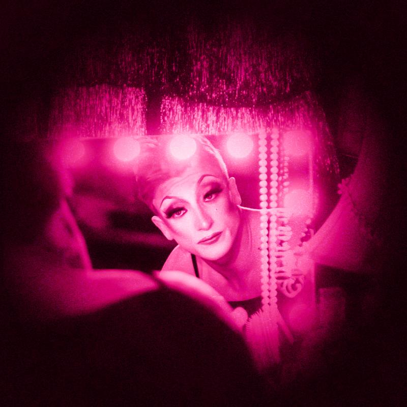 Performer stares at themselves in the mirror as they get ready. There is a neon pink filter on the photo.