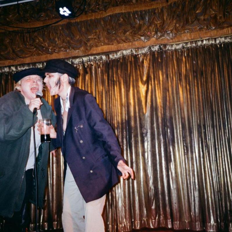 Two drag kings dressed as old men scream into a microphone on a stage with a gold curtain backdrop