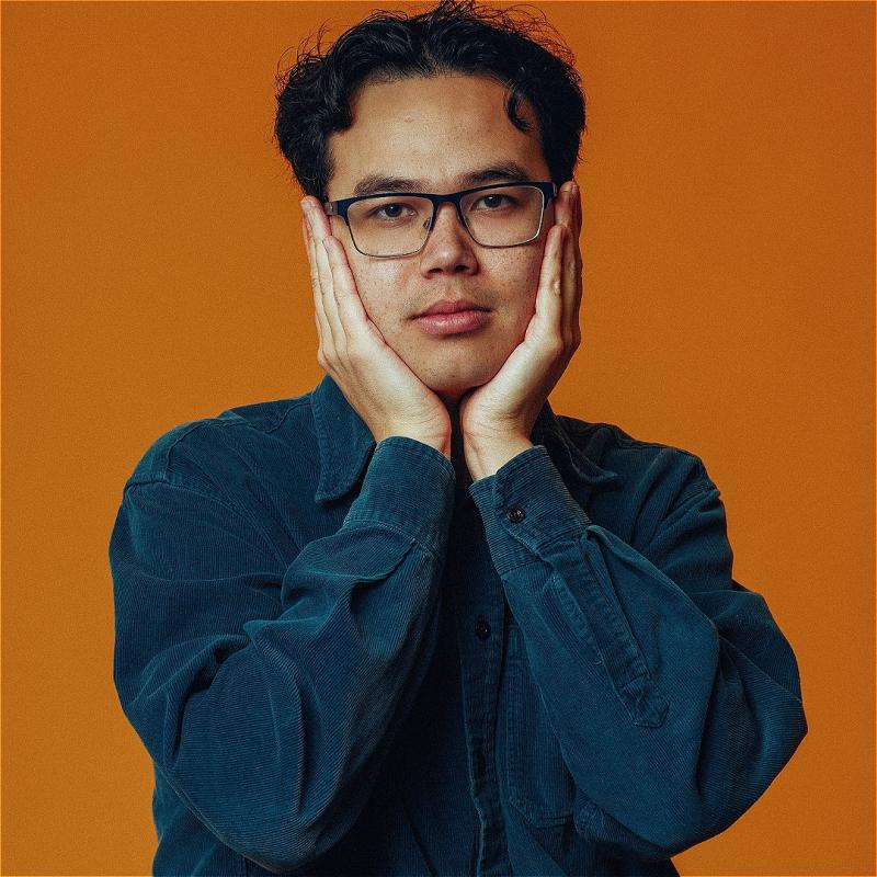 A man wearing a blue jacket standing in front of an orange background, with his head in his hands.