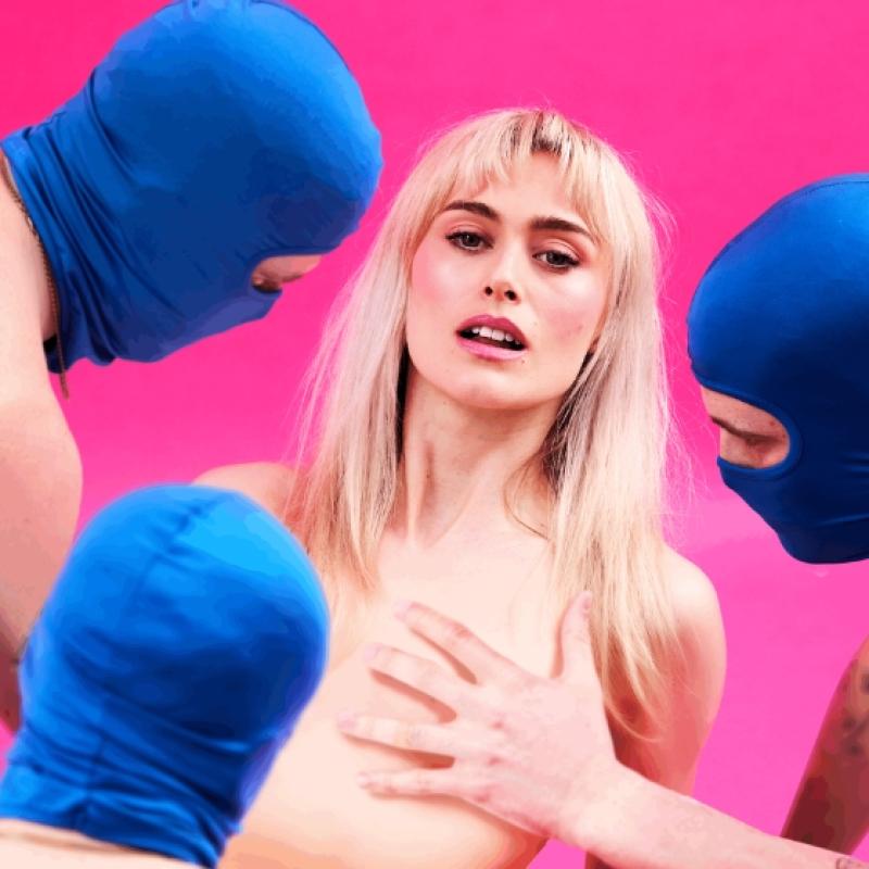 A blonde woman looks at the camera as three men in blue masks look at her