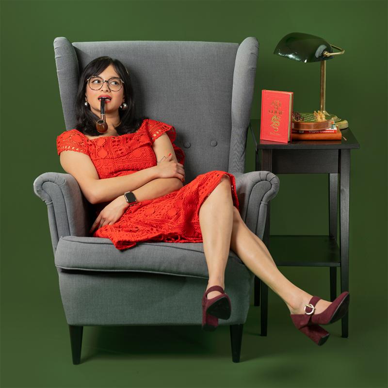 A woman wearing black glasses, a red dress, and maroon heels, sitting on a grey armchair next to a table with a red book and a lamp, in front of a green background.