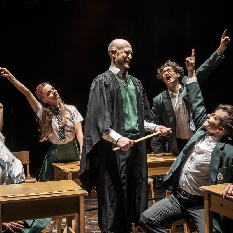 Four performers in school kids uniforms raise their arms to ask a question to a fifth performet playing the teacher