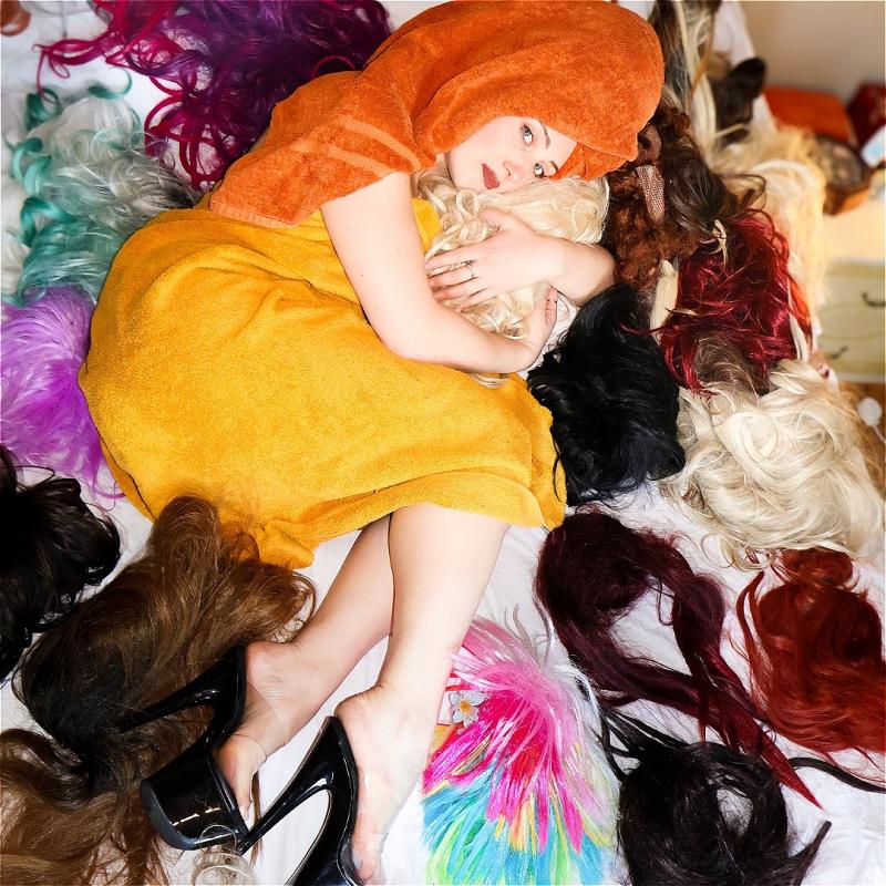 Alice lounges on a bed in the fetal position surrounded by a great many number of exuberant and vibrant wigs. She cradles one wig in her arms like a babe.