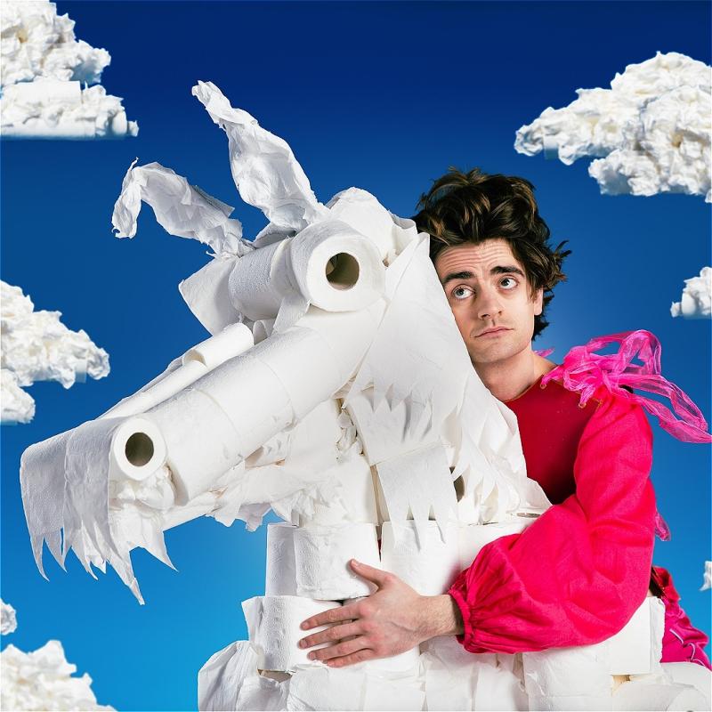 A man with very bouffant hair rides a white stallion made of toilet paper and cotton against a blue sky and fluffy clouds backdrop.