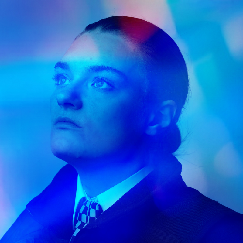 A woman dressed in a police uniform and bathed in blue light looked up and off into the distance