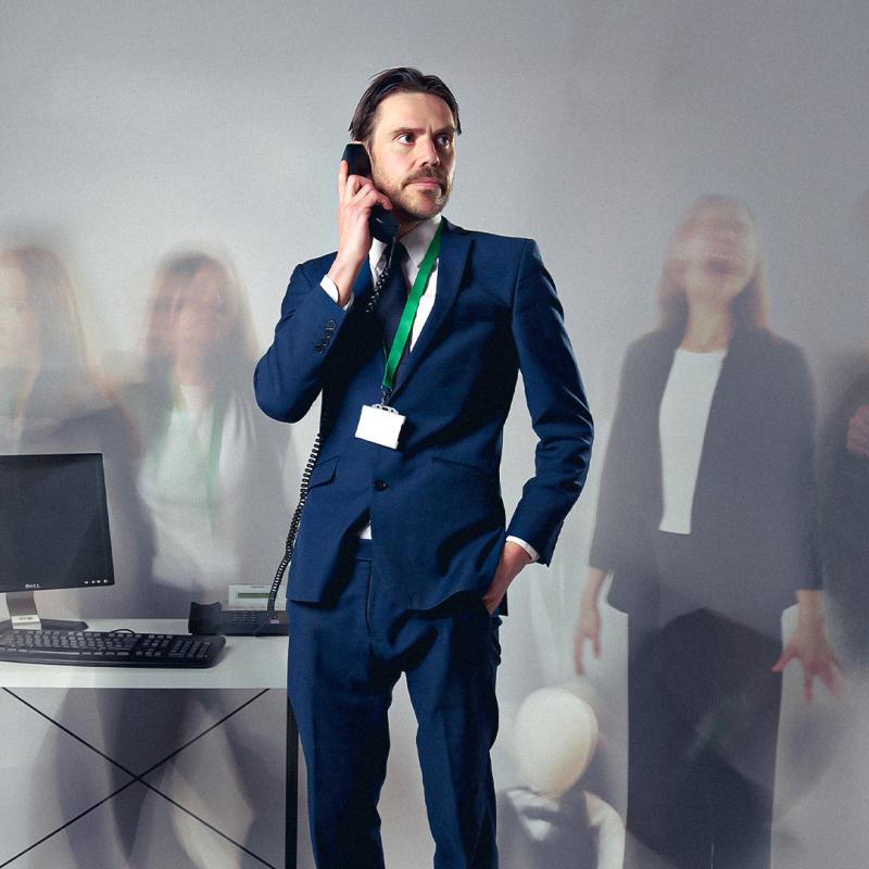 Man in suit wearing lanyard holds phone to ear looking pensively across blurry background with screaming office folk.