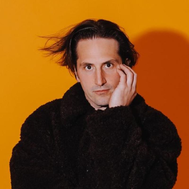 Luke Mcqueen resting his face in his hands wearing a fluffy black coat against on orange backdrop.