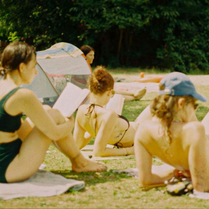 A grainy film shot of women sunbathing in a garden, some are reading.