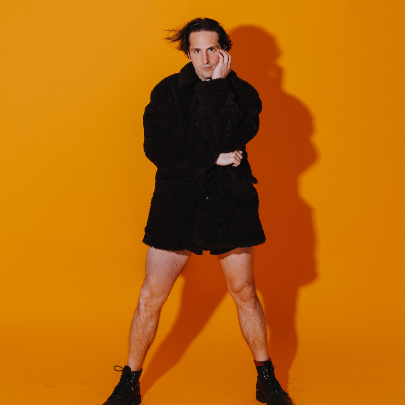 Luke Mcqueen wearing a long black raincoat and no trousers standing against a bright orange background