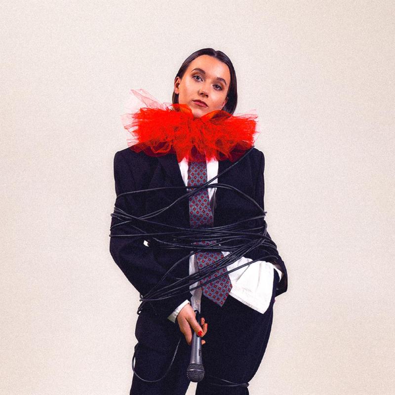Claire Noy stands in a suit, tie and large red ruff looking particularly apathetic about the fact somebody has wrapped their microphone cable around them twenty times. They hold the microphone laxly in their right hand.