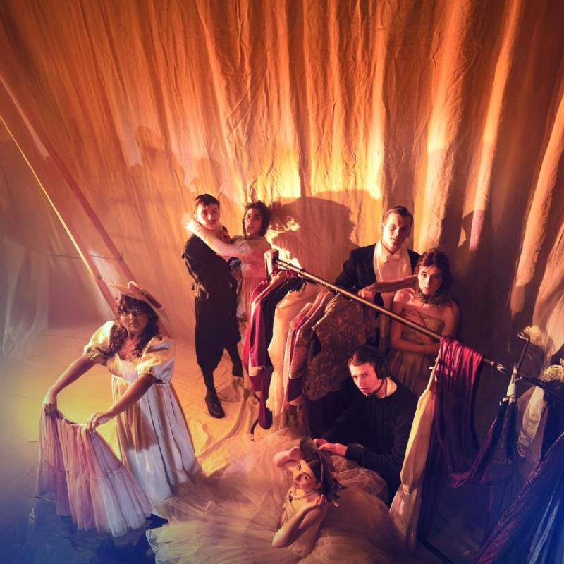 Backstage in a theatre,a group of performers in costumes look up to the camera in orange light