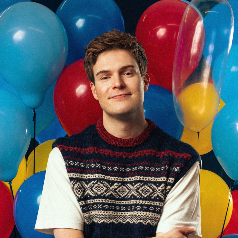 Thor stands in front of a big bunch of balloons wearing a knitted vest over a white t-shirt.