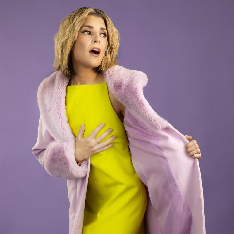 Grace Helbig wears a yellow dress and pink fur coat, looking to the side with her hand to her chest.