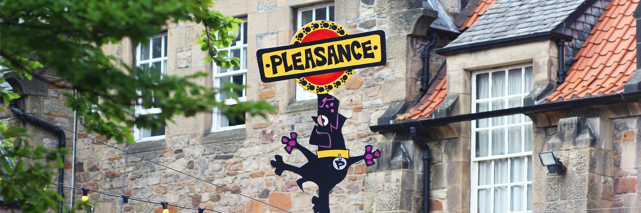 Picture of the Pleasance Courtyard in Edinburgh with the backdrop of cobbled buildings and the top of the Pleasance wayfinding pole with a cartoon black Labrador and the Pleasance logo above.