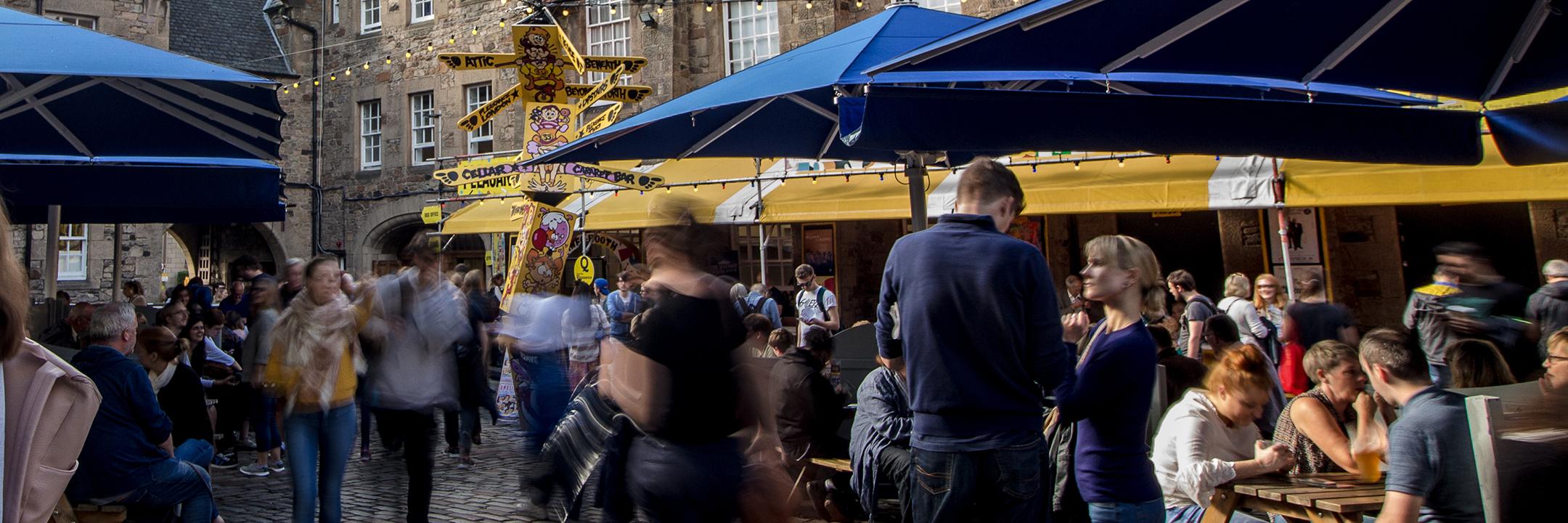 Image of the bustling Pleasance Courtyard taken from eye level. There are lots of punters standing underneath blue parasol umbrellas and yellow Pleasance signs all around.