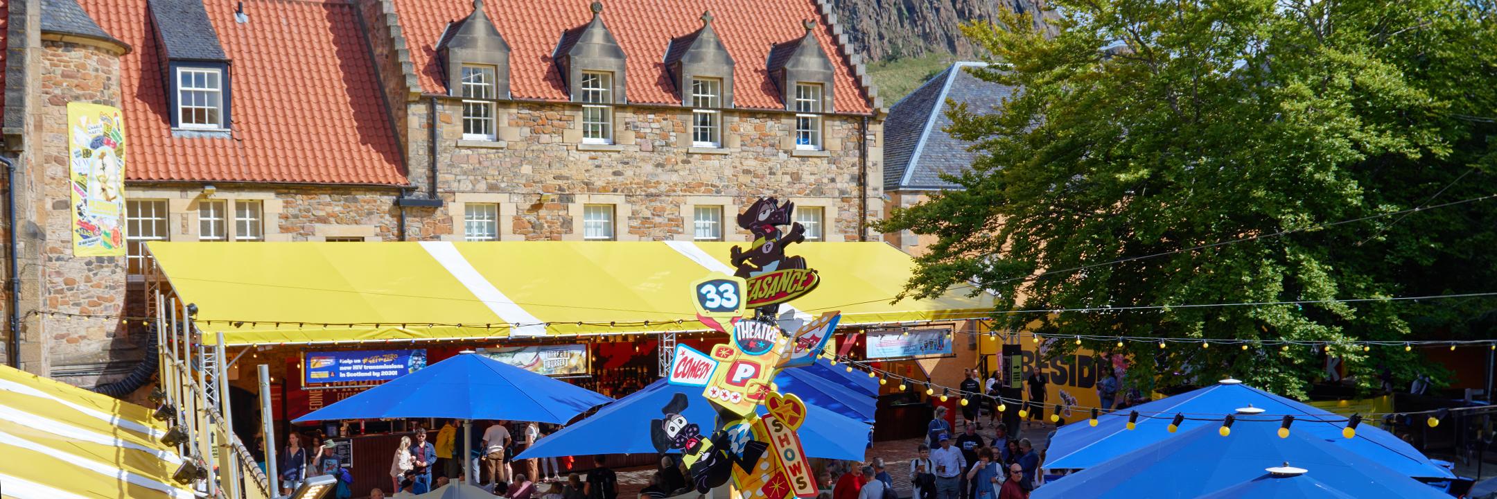 Aerial view of the Courtyard in daylight focussing on the Pleasance wayfinding pole with cartoonized examples of their programme genres and a black labrador at the top.