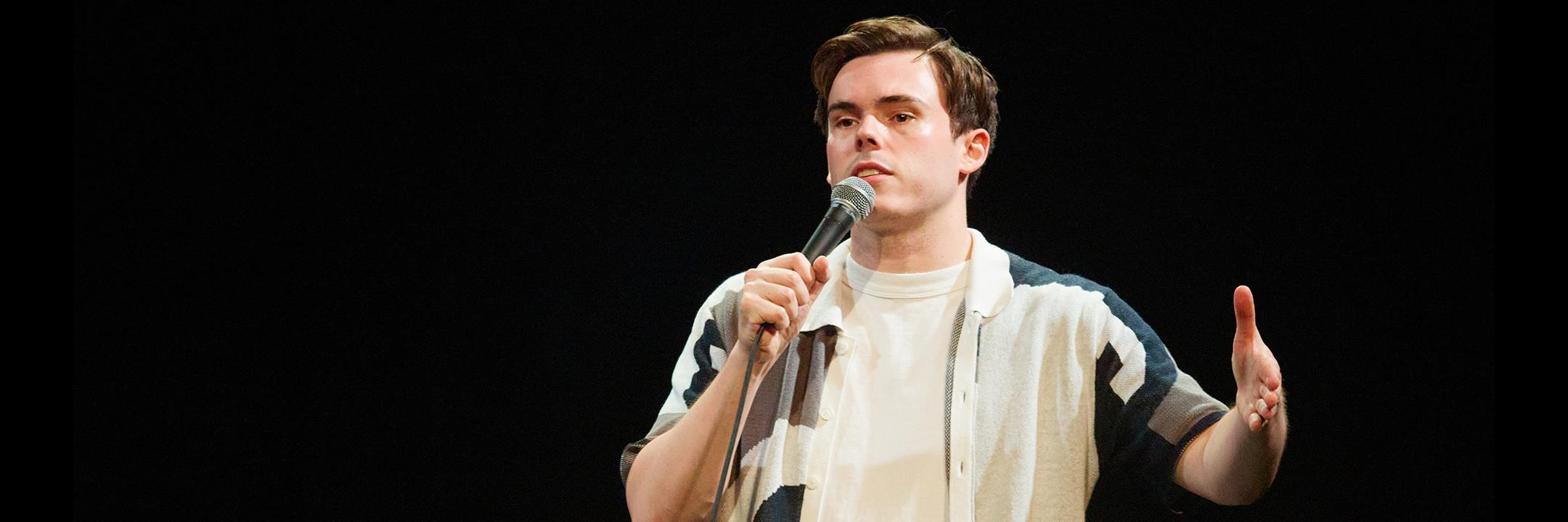 Photo of Rhys James on stage holding a microphone as they deliver a stand up set.