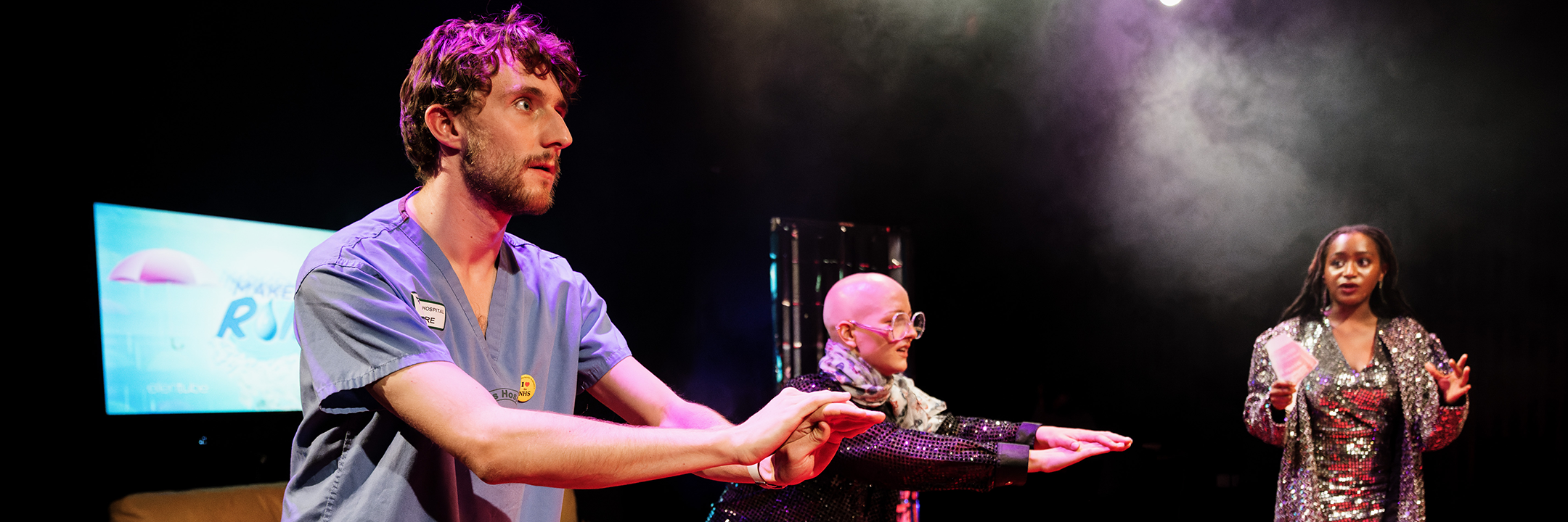 Production photo of London Associate Artists 2024-2026, Sweet Beef and their debut performance of 'I hate it here' at the Pleasance Downstairs. A trio of performers on stage - one in hospital scrubs and two in glittery clothing.