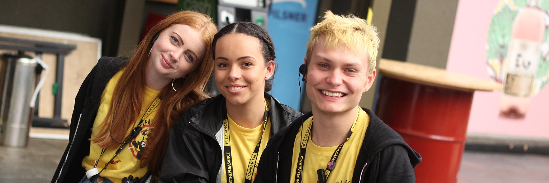A trio of smiling Pleasance volunteers wearing yellow branded t-shirts, lanyards and hoodies. They wear front of house ear pieces and carry radios at the Dome.