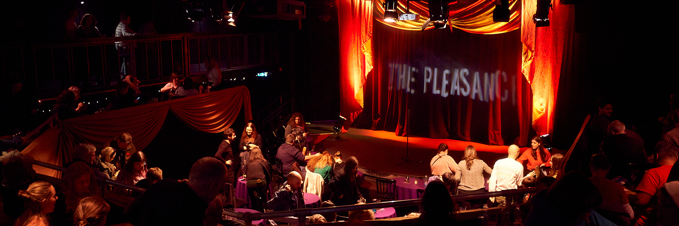 A pre-show photo of the Main House stage with The Pleasance projected onto the back wall, the auditorium is full. 