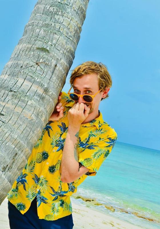 Photo of Rob standing coyly on a beach next to a palm tree with their hand partially covering their face. They are wearing a yellow Hawaiian pineapple shirt and dark blue trousers.