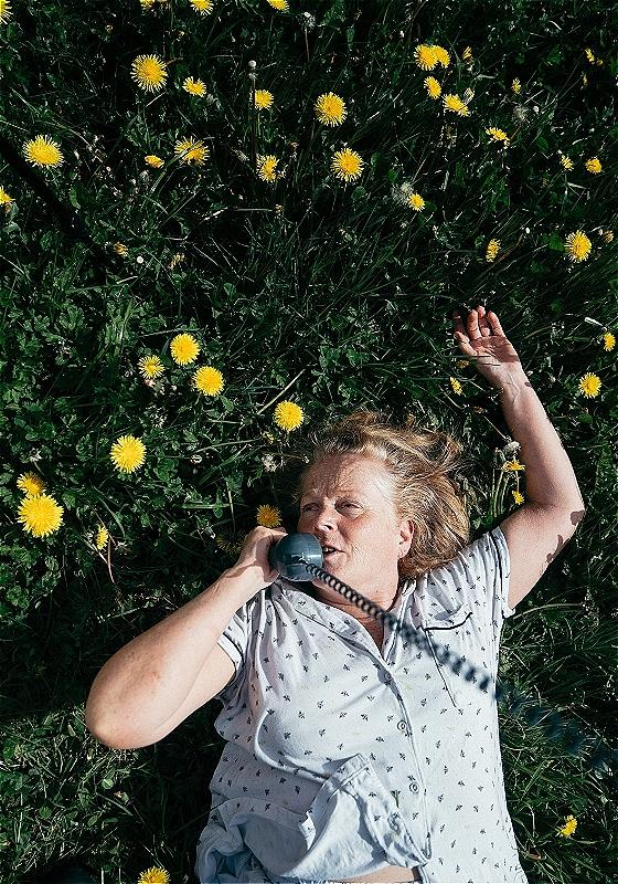 Woman lying on her back in a field of grass and dandelions, talking on an old-fashioned rotary phone.