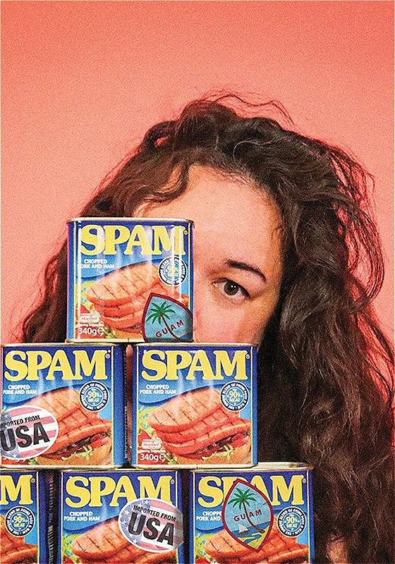 The performer is in front of a peach-coloured background, with 6 tins of Spam stacked in front of them. 