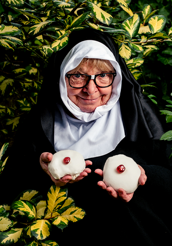 Elderly nun with glasses, smiling and holding two white skull-shaped pastries with red decorations on top, standing in front of a bush with green and yellow leaves.