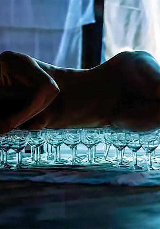 Landscape photo of a body lying across a blurred table balanced on top of wine glasses.
