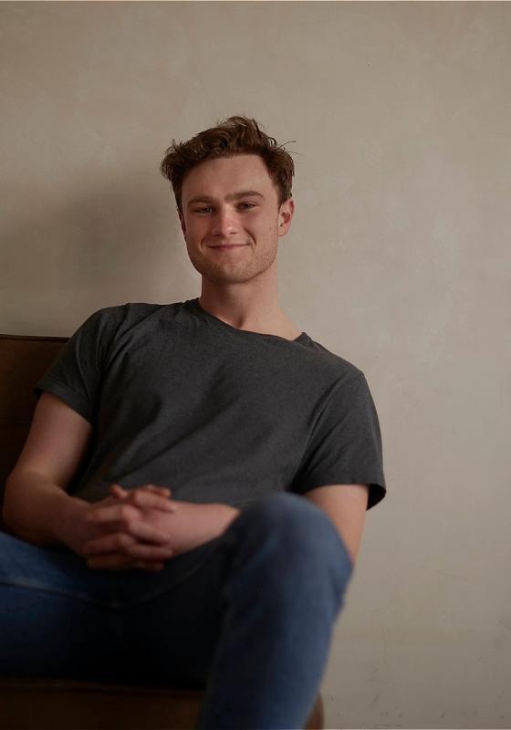 Young man with a slight smile sitting casually in a brown armchair, wearing a grey t-shirt and blue jeans, with hands clasped in front and a plain off-white wall in the background.