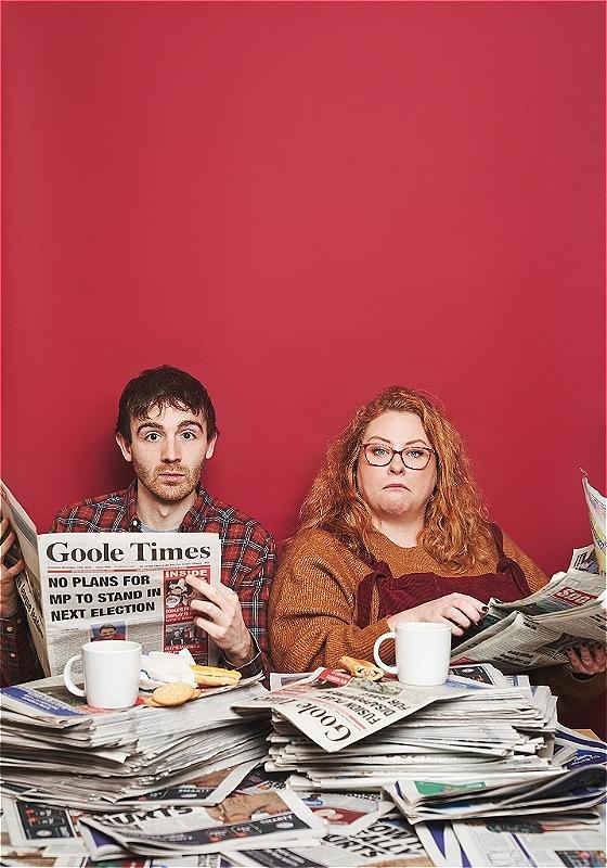 Ian and Amy sit at a table covered in newspapers with a red backdrop.