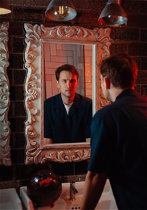 Vittorio, wearing a navy shirt, is looking inquisitively at themselves in a mirror 
