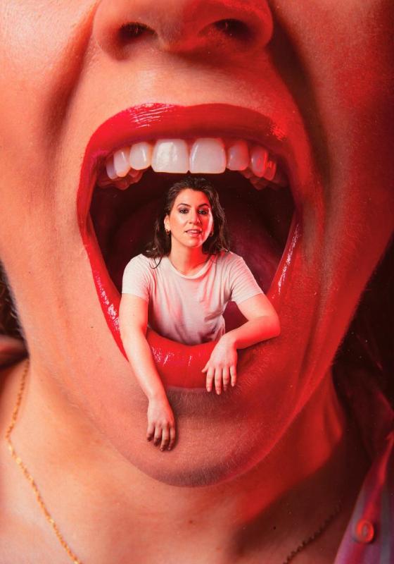 Zoe sits inside a giant open mouth with bright red lips and white teeth. She is wearing a white t-shirt and her arms hang over the bottom lip.