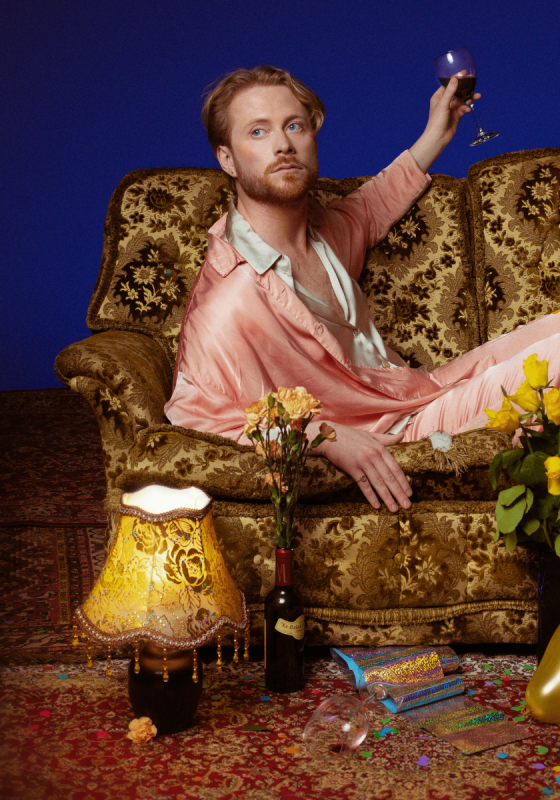 Performer sitting on gold sofa holding wine glass wearing pink silk pjs surrounded by balloons, flowers, wine and lamp