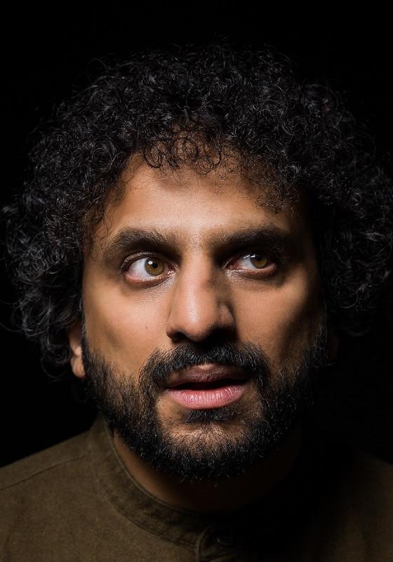 Headshot of Nish Kumar looking quizzically into the distance. He wears a brown buttonless collared shirt and the background is black.
