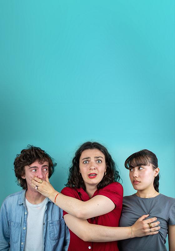 Three performers against a blue backdrop. The middle performer has her hands over the mouth of one, and holding the breast of the other.