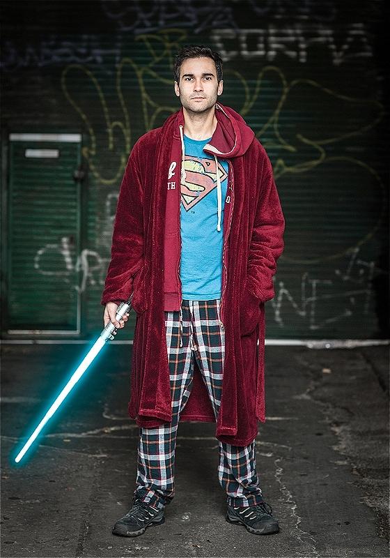 Person in a Superman shirt and plaid pajama pants holding a lightsaber, standing in front of graffiti.