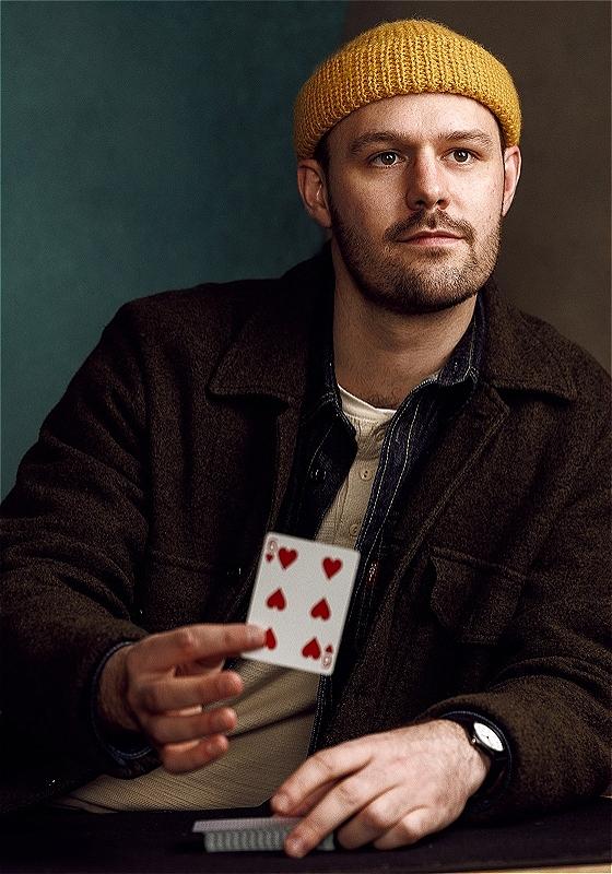 Person in brown jacket holding a playing card, sitting at a table with more cards.