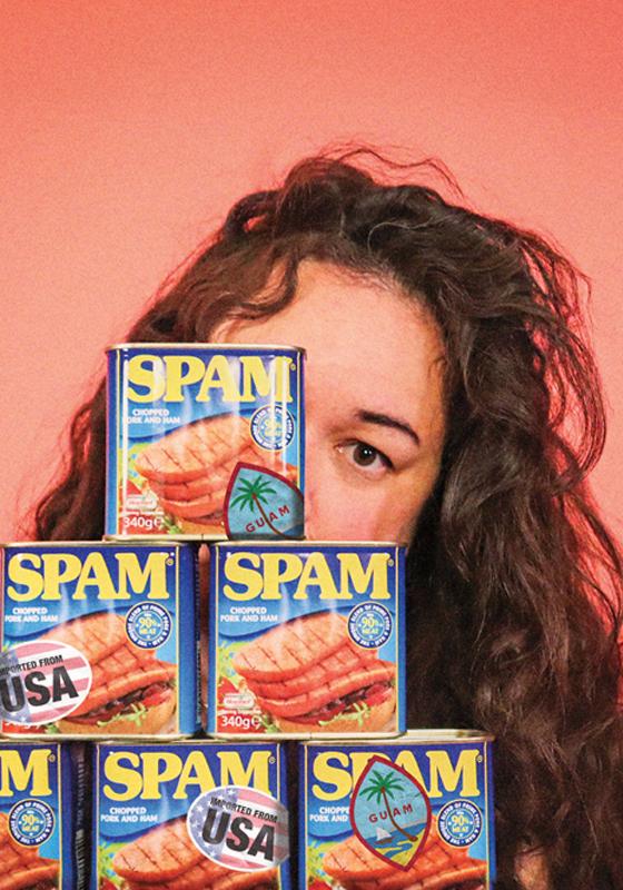 Curly-haired individual standing behind a stack of SPAM canned meat products against a coral background.