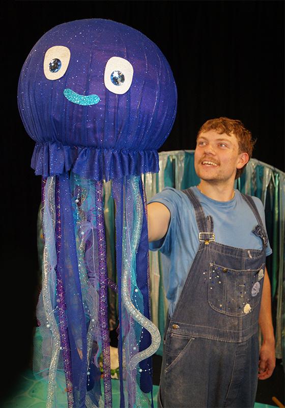 Person in overalls holding a large, decorative jellyfish costume with a sparkly blue design.