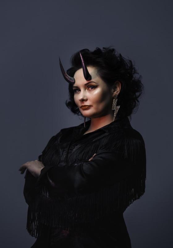 Person with black-fringed outfit, devil horns and lightning bolt earring against a grey background.